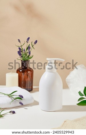 Front view of an unlabeled shower gel bottle is displayed on a table with candles, towels, lavender flowers and a brown glass vase on a pastel background. Shower gel with lavender scent. Royalty-Free Stock Photo #2405529943