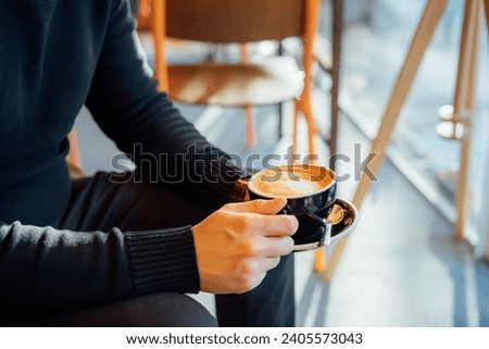 No face handsome young man in black clothes holding hot latte art or cappuccino coffee cup in modern cafe shop. Warm and cozy fall or winter moments. Take a break to relax. Soft selective focus Royalty-Free Stock Photo #2405573043