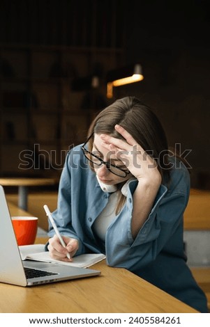Female student in casual clothes sitting at table with laptop and writing notes in notebook while doing homework in college library.