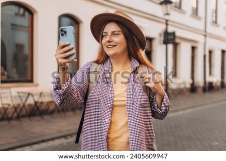 Picture of pretty young woman in hat staying on the street holding phone in hands. Redhead girl making selfie photo on urban background. Use technology concept, Traveling Europe in summer