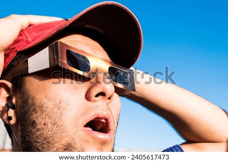 Portrait of man wearing cap and solar eclipse glasses, looking sun with blue sky at background  Royalty-Free Stock Photo #2405617473