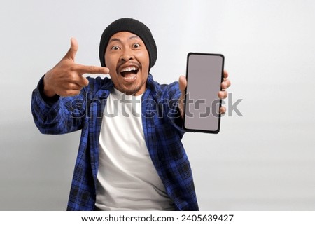 Excited Asian man, dressed in a beanie hat and casual shirt, excitedly showcases and points towards a mobile phone with an empty white screen, suggesting or recommending a new app or a special offer Royalty-Free Stock Photo #2405639427