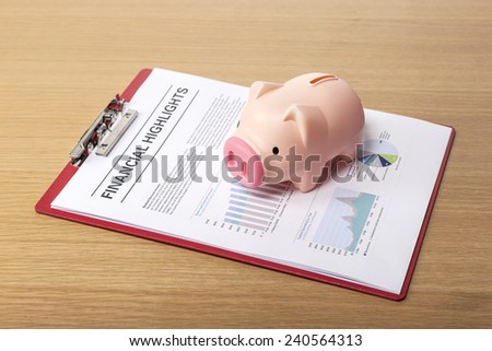 Two pig coin bank(money box), clipboard, graph paper(document) on the wooden office desk(table) behind white blind.