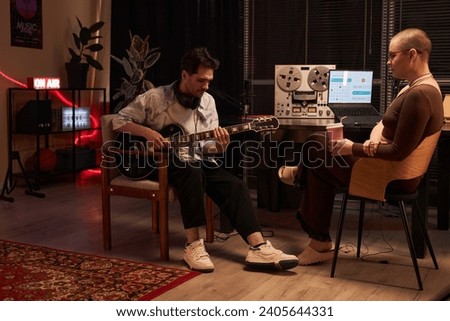 Wide shot of adult male composer with headphones on neck playing semi-acoustic guitar while sitting with female interviewer in podcast room