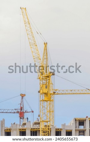 The tower cranes are working near incomplete white brick building. It is in unfinished European apartment building. it is blue sky with white clouds. It is yellow and red cranes. It is cloudy midday