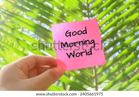 Hand holding a pink note that says GOOD MORNING WORLD with a background of trees and forest in the morning, the concept of welcoming the morning with natural views