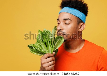 Young happy fitness trainer instructor sporty man sportsman wear orange t-shirt hold sniff green lettuce salad spend time in home gym isolated on plain yellow background. Workout sport fit abs concept Royalty-Free Stock Photo #2405666289