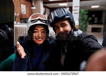 Youthful couple at luxurious ski resort, with man holding smartphone for selfie picture. Excited boyfriend and girlfriend wearing winter jacket and helmet ready for wintersport activities.