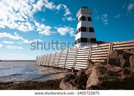 view of a tall black and white lighthouse in summer on the side of a beach with a wooden fence and rocks in the foreground Royalty-Free Stock Photo #2405722875