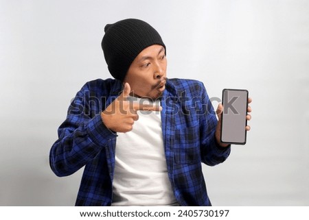 Excited Asian man, dressed in a beanie hat and casual shirt, excitedly showcases and points towards a mobile phone with an empty white screen, suggesting or recommending a new app or a special offer Royalty-Free Stock Photo #2405730197