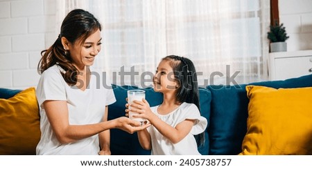 A delightful family moment as an Asian mother offers her daughter a glass of milk on the sofa. This picture radiates happiness innocence and the nutritional value of calcium.