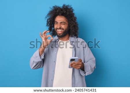 Young successful excited Arabian man holds smartphone and shows approving gesture looking at screen and winking as sign of support for new version of mobile phone stands posing in blue studio.