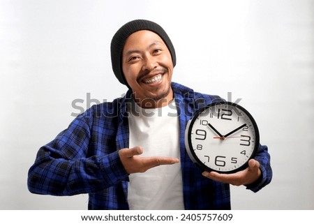 Young Asian man, dressed in a beanie hat and casual shirt, displays a clock in his hand, smiles, expressing happiness and excitement as he looks at the camera, while standing against white background