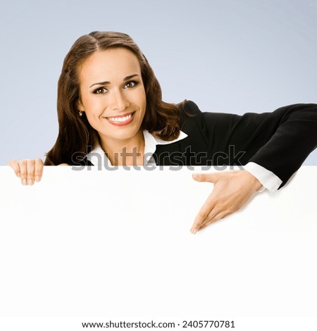 Happy smiling brunette young business woman standing behind, peeping from blank banner or mock up signboard, showing copy space for text, over grey background. Square composition