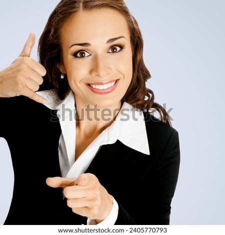 Businesswoman in black confident suit showing call me hand sign gesture and pointing at viewer, grey background. Portrait of smiling brunette woman at studio. Business concept. Square composition