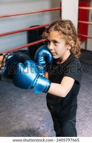 Vertical photo elementary girl, dressed in black t-shirt and tights and blue boxing gloves, learning to box in the ring with her black mid adult boxing teacher. Sport, recreation concept.