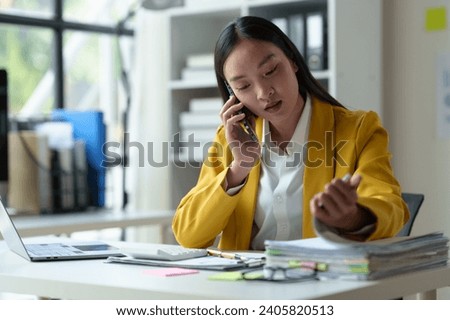 Asian businesswoman, customer sales manager of a busy company while talking on the phone with customers. Looking at documents, files, financial details, agreements, business contracts in the office.