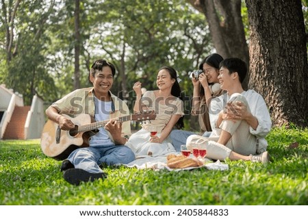 Group of cheerful diverse young Asian friends are enjoying picnicking in a beautiful green public park together, playing guitar, taking pictures, enjoying food and chatting. Friendship and lifestyle