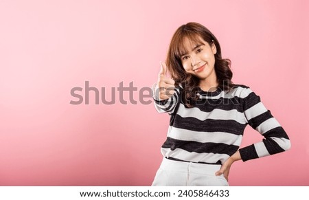 A smiling Asian woman in a studio shot on pink background confidently gives a thumbs-up 'Ok' sign, symbolizing agreement and success. Her cheerful expression radiates positivity and satisfaction.