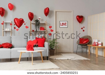 Interior of festive living room with grey sofa, heart-shaped balloons and glasses of wine on coffee table. Valentine's Day celebration Royalty-Free Stock Photo #2405850251