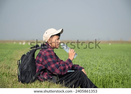 Asian boy in plaid shirt wears cap and has a backpack, holding a drinking bottle, sitting on ridge rice field to observe pm. 2.5 smoke and birds on rice fields, soft focus, new edited.