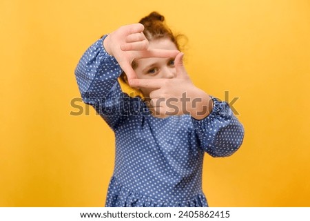Portrait of cute little girl making frame with fingers, looking at camera, pretending to make photography, posing isolated on yellow color background wall in studio with copy space. Selective focus