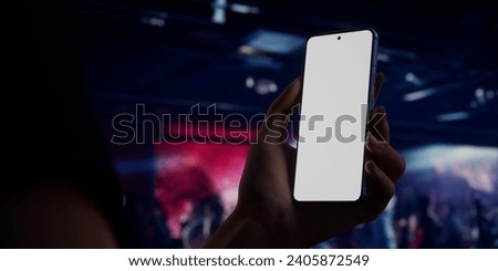 Caucasian woman holding a phone in two hands, people dancing in the night club in the background. Blank white screen smartphone mockup. Horizontal orientation