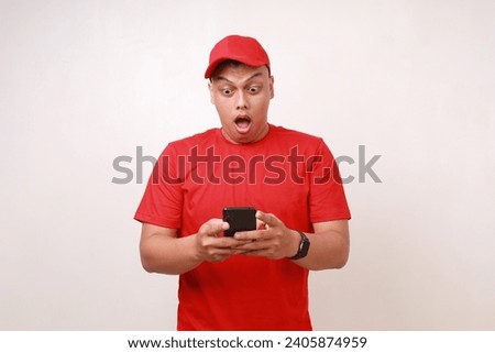 Shocked Asian delivery guy employee man in red standing while holding a cell phone. Isolated on white