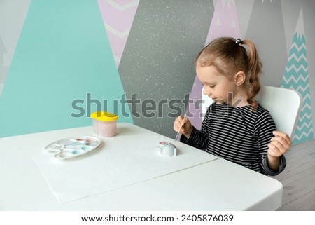 A little curly girl of European appearance of 3-4 years old sits at the desk and paints a plaster figure of a hippo. Education concept. Home hobby and fun game. Royalty-Free Stock Photo #2405876039