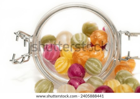 A close-up view of a serving jar with colourful sugar balls. The sugar balls roll out of a jar with a lid. The background is light-coloured.