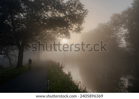 A solitary cyclist embarks on a journey along a riverside path shrouded in mist in this evocative image. The morning sun, a muted sphere, pierces through the fog, casting a gentle glow on the scene Royalty-Free Stock Photo #2405896369