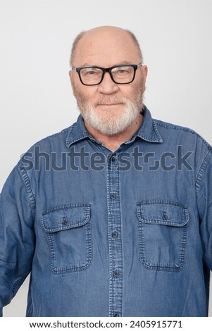 Portrait of an attractive gray-haired man with glasses wearing a denim shirt isolated on a gray background. Bearded grandfather of the European type.