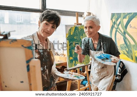 mature female artists with palettes looking at easel in art workshop, creativity and collaboration Royalty-Free Stock Photo #2405921727