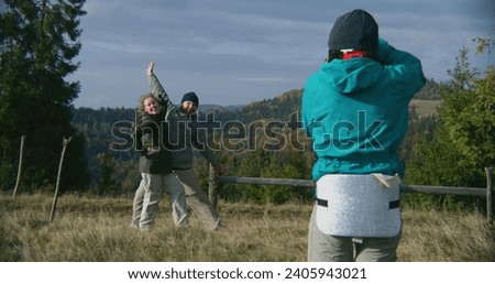 Young hiker takes pictures of Caucasian tourist family using camera. Happy couple of travelers posing on top of hill against beautiful landscape during hiking trip in mountains. Outdoor enthusiasts.