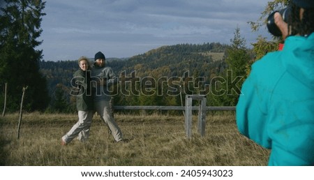 Young hiker takes pictures of Caucasian tourist family using camera. Happy couple of travelers posing on top of hill against beautiful landscape during hiking trip in mountains. Outdoor enthusiasts.