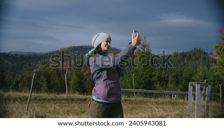 Female hiker takes selfie using mobile phone standing in front of beautiful scenery on the hill. Caucasian woman holds cup of tea and looks at smartphone. Tourist during trip or trek in the mountains.