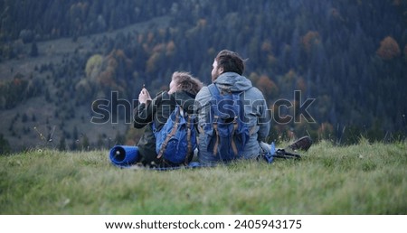 Happy Caucasian couple of hikers sits on the hill, looks at photos on the phone or surfs the Internet. Family of travelers during hiking trip or trek in the mountains. Tourism and outdoor recreation.
