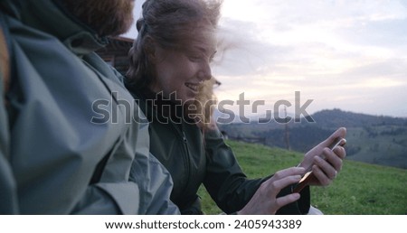 Happy Caucasian couple of travelers sits on the hill, looks at pictures on the phone or surfs the Internet. Tourist family during hike or trekking trip in the mountains. Tourism and travel concept.