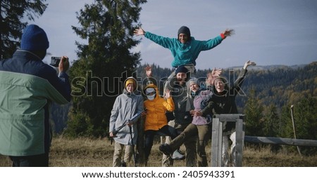 African American man takes group pictures of his multiethnic friends against beautiful mountains landscape during hiking trip or trek. Diverse tourists smile and wave at camera. Outdoor enthusiasts.