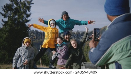African American man takes group pictures of his multiethnic friends against beautiful mountains landscape during hiking trip or trek. Diverse tourists smile and wave at camera. Outdoor enthusiasts.