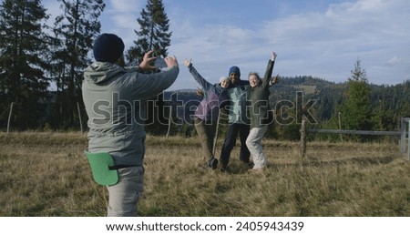 Man takes group picture of multiethnic friends or hiking buddies on the top of the hill against beautiful scenery using camera. Diverse hikers during trip or hike in mountains. Outdoor enthusiasts.