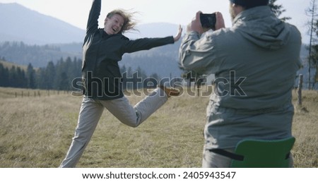Caucasian man takes pictures of his wife on hilltop against scenic nature view using professional camera. Couple of travelers, tourist family during hiking trip or trek in mountains. Active leisure.
