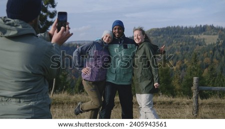 Man takes group picture of multiethnic friends or hiking buddies on the top of the hill against beautiful scenery using camera. Diverse hikers during trip or hike in mountains. Outdoor enthusiasts.