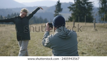 Caucasian man takes pictures of his wife on hilltop against scenic nature view using professional camera. Couple of travelers, tourist family during hiking trip or trek in mountains. Active leisure.
