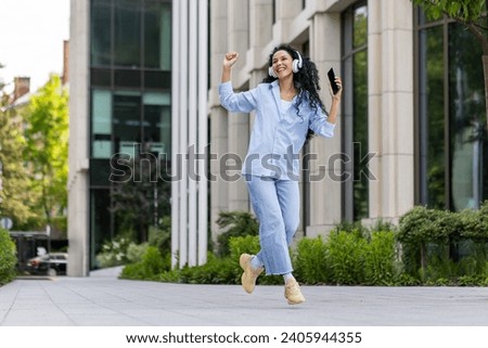 Young joyful beautiful woman dancing and singing in headphones while walking in the city, Hispanic woman with curly hair uses an application on the phone, to listen to music online. Royalty-Free Stock Photo #2405944355