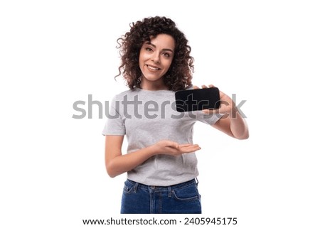 young funny pretty brunette curly caucasian lady in casual jeans and a t-shirt demonstrates a smartphone screen with a mockup for advertising on a white background with copy space