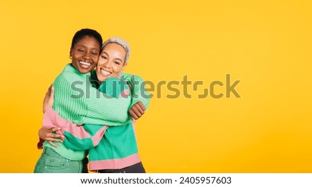 Two young cheerful women wearing green clothes embracing in a studio with a yellow background. Royalty-Free Stock Photo #2405957603