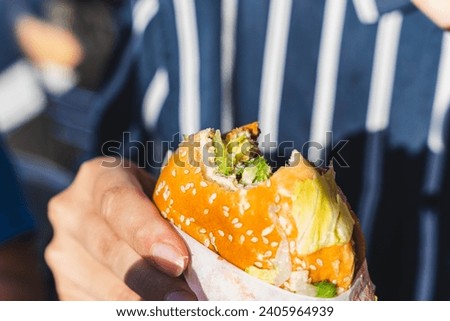 A close-up shot of a girl's hand holding a delicious fresh burger with a bite taken, garnished with herbs and sesame seeds. The juicy patty is visible, creating an enticing and mouth-watering image.