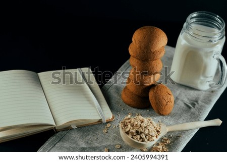 Stack of fresh oatmeal cookies with milk in jar, raw oat flakes in large wooden spoon, open notebook on black background. Still-life Royalty-Free Stock Photo #2405994391