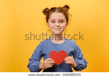 Portrait of pretty smiling preteen girl holding paper heart, happy looking at camera, posing isolated over yellow color background wall in studio. Greeting card on Valentine's day, 14 February concept